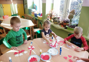 Grupa dzieci przy stolikach wykonuje z papierowych serduszek biało - czerwone kotyliony.